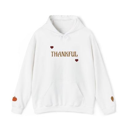 A white pullover hoodie with 'THANKFUL' printed in brown across the chest, flanked by two small brown hearts. The hoodie features a front kangaroo pocket, white drawstrings, a small orange pumpkin graphic on the left sleeve, and a small brown leaf graphic on the right sleeve.