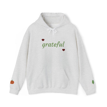 A light heather gray pullover hoodie with 'grateful' written in green script across the chest, accented by two small red hearts. The hoodie features a white drawstring hood, a front kangaroo pocket, a small orange pumpkin graphic on the left sleeve, and a small green leaf graphic on the right sleeve.
