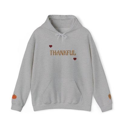 A light gray heather pullover hoodie with 'THANKFUL' printed in brown across the chest, flanked by two small red hearts. The hoodie features a white drawstring hood, a front kangaroo pocket, and small autumn-themed graphics on the cuffs, including a small orange pumpkin on the left sleeve and a leaf on the right sleeve.