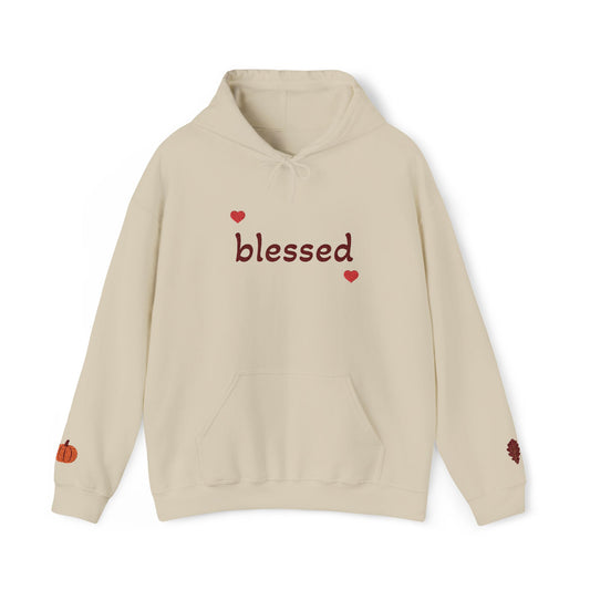 A beige pullover hoodie with 'blessed' written in dark red script across the chest, flanked by two small red hearts. The hoodie features a drawstring hood, a front kangaroo pocket, a small orange pumpkin graphic on the left sleeve, and a small dark red berry graphic on the right sleeve.