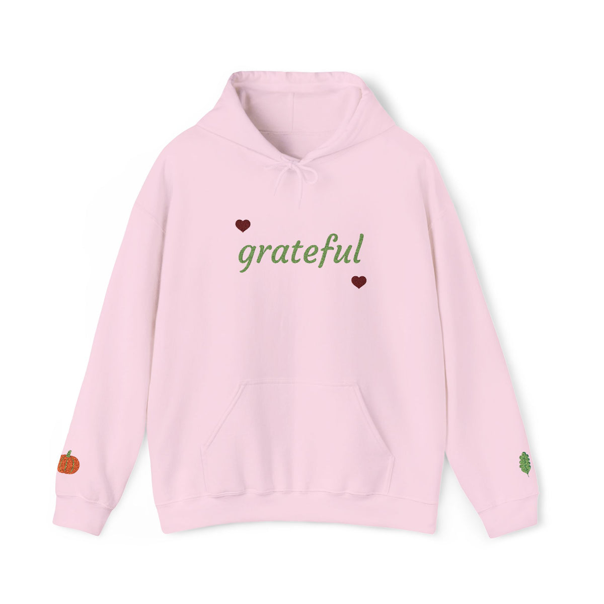 A light pink pullover hoodie with 'grateful' written in green script across the chest, flanked by two small dark red hearts. The hoodie features a front kangaroo pocket and small decorative graphics on the cuffs, including a small orange pumpkin on the left sleeve and a small green leaf on the right sleeve.