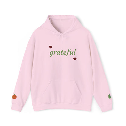 A light pink pullover hoodie with 'grateful' written in green script across the chest, flanked by two small dark red hearts. The hoodie features a front kangaroo pocket and small decorative graphics on the cuffs, including a small orange pumpkin on the left sleeve and a small green leaf on the right sleeve.