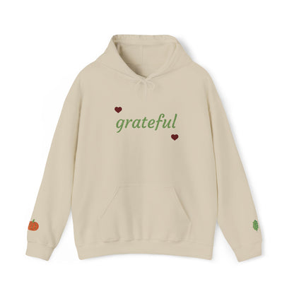 A beige pullover hoodie with 'grateful' written in green script across the chest, flanked by two small red hearts. The hoodie features a drawstring hood, a front kangaroo pocket, a small orange pumpkin graphic on the left sleeve, and a small green leaf graphic on the right sleeve.