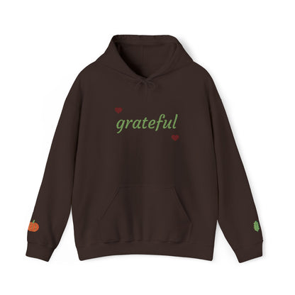 A dark brown pullover hoodie with 'grateful' written in green script across the chest, flanked by two small red hearts. The hoodie features a drawstring hood, a front kangaroo pocket, a small orange pumpkin graphic on the right sleeve, and a small green leaf graphic on the left sleeve, shown against a plain white background.