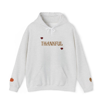 A light heather gray pullover hoodie with 'THANKFUL' printed in brown across the chest, flanked by two small brown hearts. The hoodie features a white drawstring hood, a front kangaroo pocket, a small orange pumpkin graphic on the left sleeve, and a small brown leaf graphic on the right sleeve, shown against a plain white background.