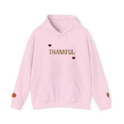 A light pink pullover hoodie with 'THANKFUL' printed in brown across the chest, flanked by two small brown hearts. The hoodie features a drawstring hood, a front kangaroo pocket, and small autumn-themed graphics on the cuffs, shown against a plain white background.